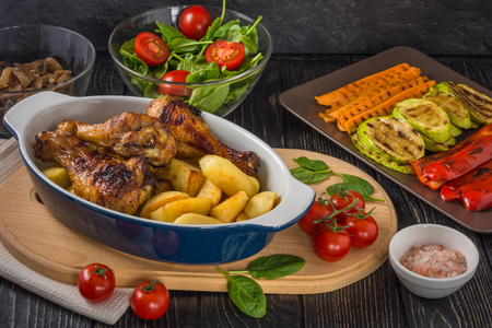 Tasty and nutritious meal with baked chicken drumsticks and potato in ceramic dish,  grilled vegetables, salad and tomatoes on the background of dark tableの写真素材