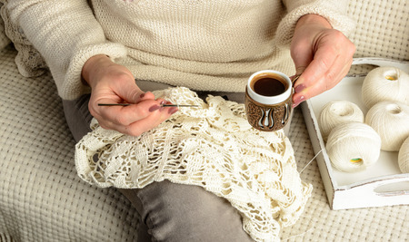 woman sitting on sofa covered with white woolen blanket, crochet tablecloth and drinking hot black coffeeの写真素材