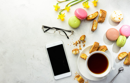 Cup of black tea with tasty almond cookies, rich in vitamins, minerals, varicolored macaroons, smartphone, glasses and flowers on a white background in light key. Top view , copy space.の写真素材