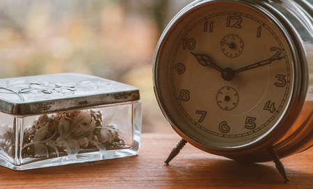 Vintage metal analog alarm clock with Arabic numbers and wind-up mechanism, sitting on a wooden bench with retro background. Time is now, time is money, old times concept. Copypace included.の写真素材