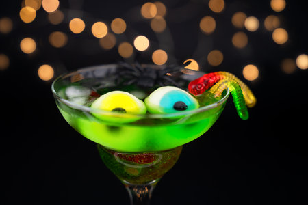Spooky green Halloween cocktail with eyes, black spider and candy worms in a glass and a garland in the background. Festive party. Close-up. Selective focus.の写真素材