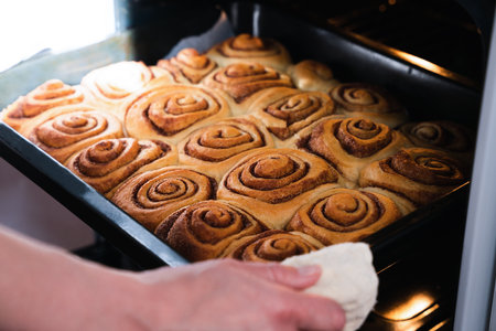 A woman takes out fresh appetizing homemade buns with cinnamon from the oven. Homemade baking. Traditional American dessert - cinnabon buns. Close-up. Selective focus.の写真素材