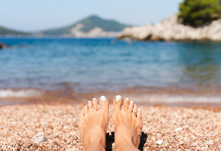 Feet of young woman with pedicure on a pebble beach on vacation. Enjoying the sun on a summer sunny day at the sea. Vacation and travel concept. Close-up.の写真素材
