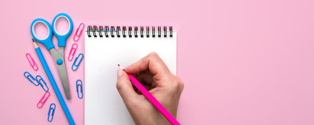 A woman's hand writes with a pink pencil in an empty notebook. Banner. Pink background. Flatlay composition. Top view. Copy space.の写真素材