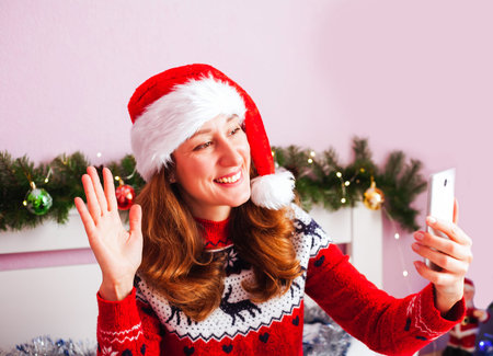 Smiling young woman in Santa hat waving her hand and calling to friends or parents by smartphone....