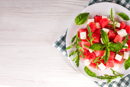 Fresh salad with watermelon, feta cheese, arugula and basil leaves on plate on the wooden white background. Healthy food concept. Close-up. Top view. Place for text.の写真素材