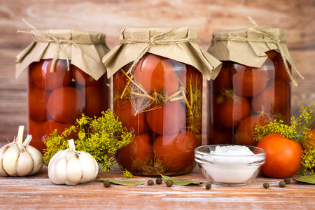 Pickled homemade tomatoes with garlic, dill, and spices in jars on the wooden table at the kitchen. Autumn food storage. Close-up.の写真素材