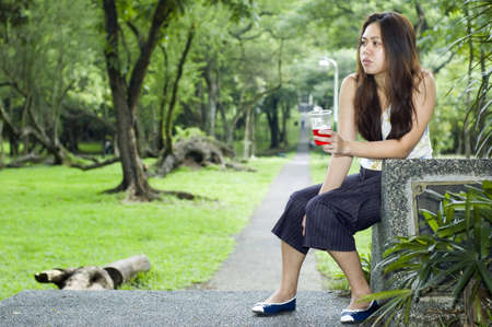 A tired looking woman waiting along a long path while holding a cup of red drink  on her handの写真素材