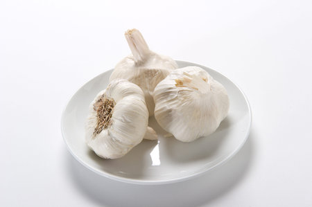 Three bulbs of fresh, white garlic on a white plateの写真素材
