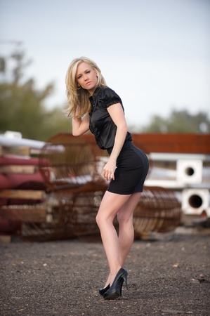 Beautiful blond fashion model with her hands on her hip and her head tilted up, wearing a short black skirt posing at a construction site outdoors.の写真素材