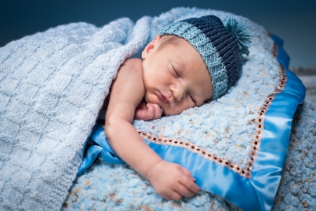 Newborn boy asleep on soft blue blankets, wearing a blue knit hat.の写真素材