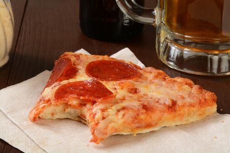 A slice of pepperoni pizza with a bite out of it and a mug of beerの写真素材