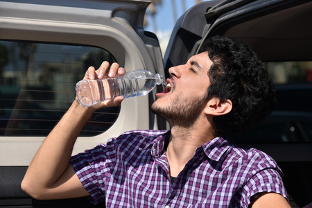 Handsome young man pouring water into his mouth on the tailgate of his carの写真素材