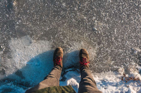 A man stands on frozen Lake with transparent ice with deep cracks. Beautiful winter landscape with clean, smooth. Top view of the legs with winter shoesの写真素材
