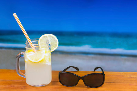 Refreshing lemon drink on a wooden table with sunglasses against a out of focus beach and ocean...