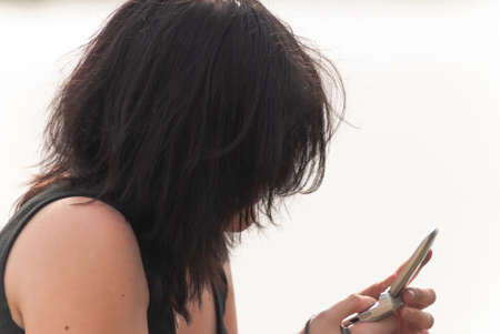 Punk emo girl with long black hair and eyeliner, young adult using her mobile phone while sitting outdoors at the lake, holding a camera in her right hand, horizontalの写真素材