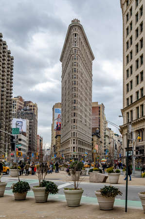 Flatiron Building New York City during daytime with overcast, lots of people, traffic, yellow taxi cabs and plants in forefront, view from low angle, verticalのeditorial素材