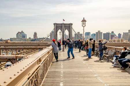 Brooklyn Bridge New York City in the evening during winter with tourists walking, taking photos and relaxing on benches on the bridge, wide angle shot, skyscraper in the background, horizontalのeditorial素材