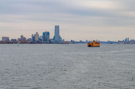 Yellow Staten Island Ferry on Hudson river in front of Jersey City in the evening, New York, during winter with overcast, wide angle shot, horizontalのeditorial素材
