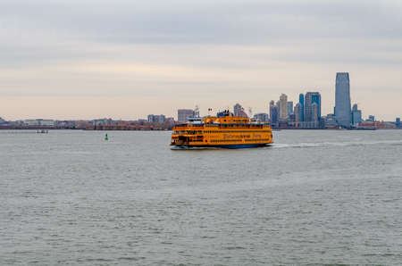 Yellow Staten Island Ferry on Hudson river passing by in front of Jersey City with office building skyscraper, New York, during winter evening with overcast, horizontalのeditorial素材