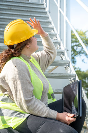 Engineer woman with yellow helmet and brown curly hair is looking up while sitting on the stairs of a wind turbine with a digital tabletの写真素材