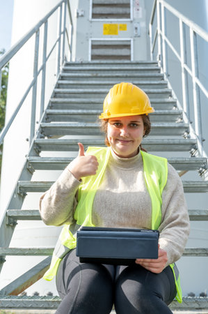 Female engineer with brown curly hair and yellow helmet is sitting on the stairs in front of the entrance of a wind turbine, working on a digital tablet, thumbs up, smiling and looking at cameraの写真素材