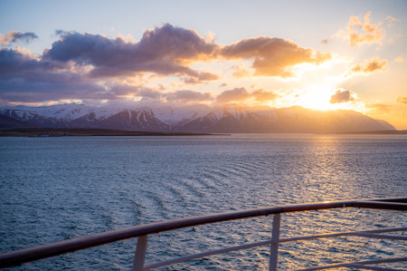 Beautiful sunset above snow mountains near Seydisfjordur, Iceland with cruise ship railing and sea in front, clouds above the mountain, wide angle shotの写真素材