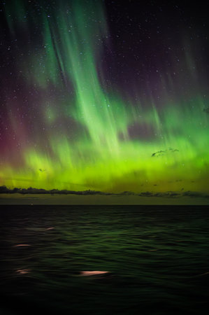 A mesmerizing display of the Northern Lights illuminates the night sky over a serene ocean, casting a vibrant green and purple glow across the waterの写真素材