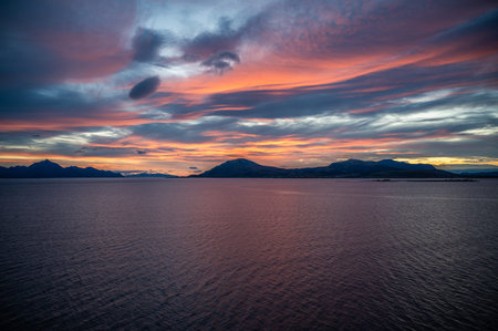 A serene sunset over a vast body of water with dramatic clouds and distant mountains, casting a warm...