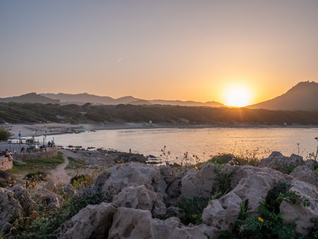 A serene sunset at Cala Rajada, Mallorca over a calm body of water with a rocky shoreline and a distant mountain range, bathed in warm hues, Majorcaの写真素材