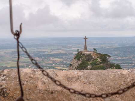 Santuari de Sant Salvador, Mallorca, large cross stands atop a rocky hill, overlooking a vast landscape with fields and distant hills under a cloudy sky.の写真素材