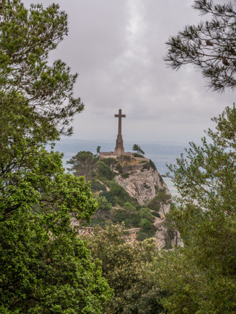 Santuari de Sant Salvador, Mallorca, large cross stands atop a rocky hill surrounded by lush green trees, overlooking a distant landscape under a cloudy sky, Majorcaの写真素材