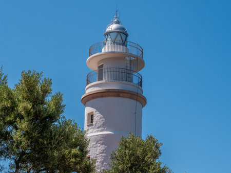 Far del Cap Gros, Port de Soller, Mallorca white lighthouse with a metal railing stands against a clear blue sky, surrounded by lush green trees, Majorcaの写真素材