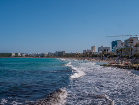 Cala Millor, Mallorca, bustling beach with clear blue waters and a city skyline in the background under a bright, sunny skyの写真素材