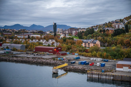 Narvik, Norway, scenic harbor town with autumn foliage, featuring industrial buildings and a waterfront with a dock and construction equipment. Mountains and modern buildings rise in the backgroundの写真素材