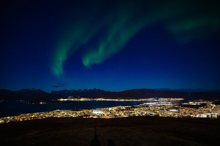 A breathtaking night view of Tromso, Norway with the aurora borealis illuminating the sky, set against a backdrop of mountains and a calm body of water, view from Storsteinenの写真素材