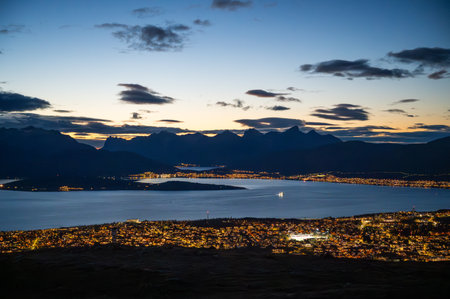 A stunning cityscape of Tromso, Norway with illuminated buildings overlooks a serene lake at twilight, with majestic mountains in the background, at the top of Storsteinenの写真素材