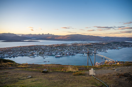 A panoramic view of Tromso, Norway with a harbor, surrounded by mountains and a bridge, set against a serene sky at sunsetの写真素材