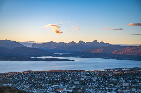 A panoramic view of Tromso, Norway, nestled between a serene lake and majestic mountains, bathed in...