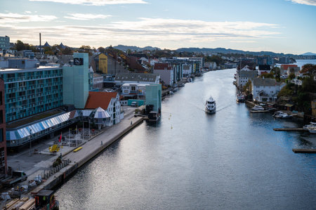 Haugesund scenic harbor with a boat sailing down a calm waterway lined with modern and traditional buildings, set against a backdrop of hills and a clear sky, Norwayの写真素材