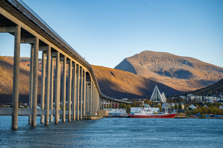 Tromsobrua Bridge, spans across a body of water, with a red and white boat docked near a mountainous...
