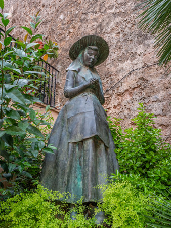 A bronze statue of a woman in traditional attire stands amidst lush greenery, with a stone wall in the background, Valldemossa, Mallorcaの写真素材