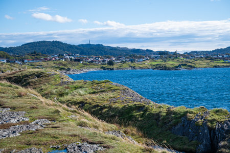 Kyststien, Haugesund, Norway, coastal scene with a serene blue sea, green hills, and a quaint village nestled along the shoreline under a partly cloudy skyの写真素材