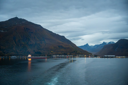 Narvik, Norway, serene harbor scene with a ship sailing under a cloudy sky, surrounded by rugged mountains and a distant townの写真素材