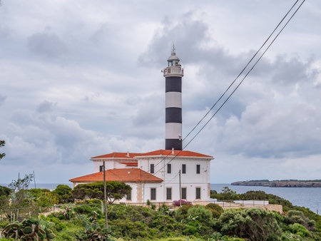 Portocolom lighthouse, Mallorca, lighthouse stands on a coastal island, surrounded by lush greenery and a serene sea under a cloudy skyの写真素材