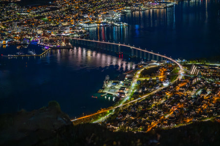 Tromso, Norway, stunning night view of the vibrant city by the water, illuminated by lights reflecting off the calm sea and a long bridgeの写真素材