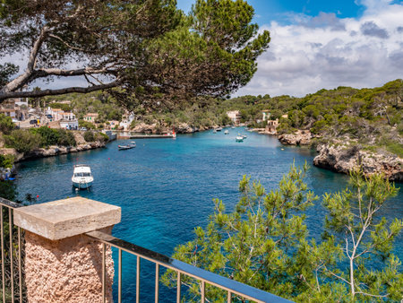 Cala Figuera, Mallorca, coastal scene with clear blue waters, lush greenery, and a small harbor filled with boats, framed by a stone railing and a pine tree, Majorcaの写真素材
