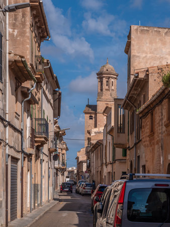 Llucmajor, Mallorca, narrow street lined with old buildings and parked cars stretches into the distance, with a historic tower visible in the background under a clear blue sky, Majorcaの写真素材
