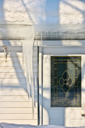 Large icicles and ice dams form at the edge of the snow-covered roof above frozen eavestrough gutterの写真素材