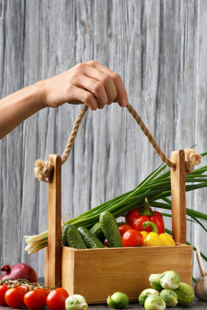 Tomatoes, cucumbers, peppers, Brussels sprouts, onions and garlic in a wooden basket. Basket with vegetables in hands on a wooden background. Useful vegetables. Proper nutrition. Vegan food.の写真素材